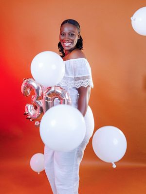 A smiling woman in a white dress holds silver 30 balloons and white balloons against an orange background.