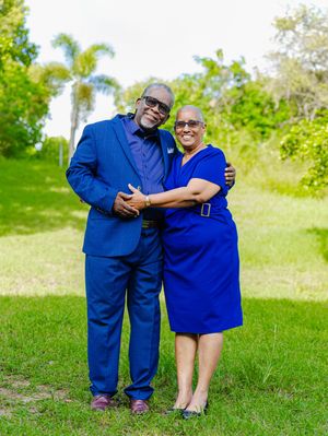 Elegant couple in matching royal blue outfits posing together on green grass with palm trees in background.