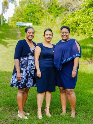 Three women in navy blue dresses smiling together outdoors on green grass.