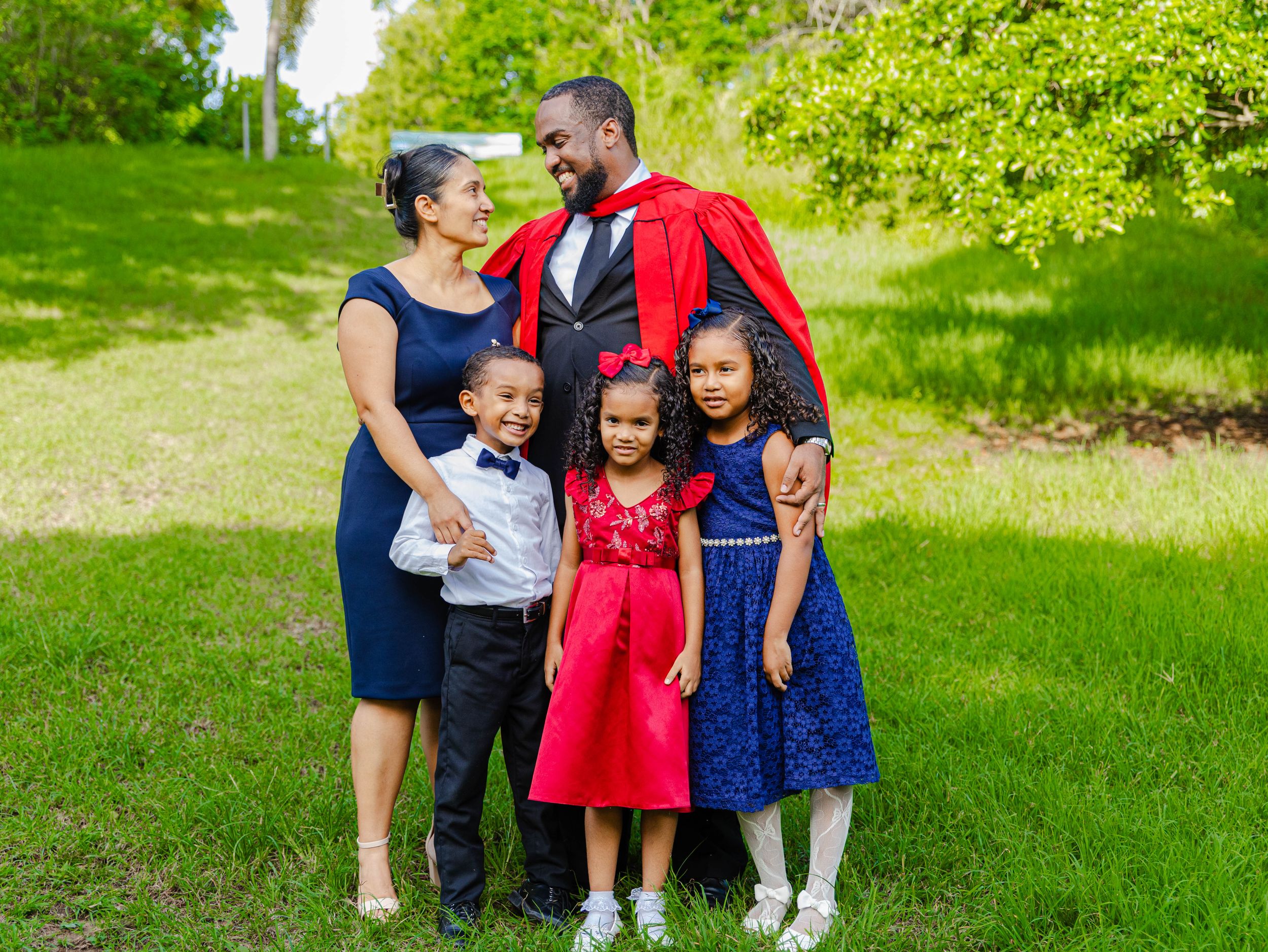 Family of five celebrating graduation with man in red academic gown surrounded by wife and three children outdoors.