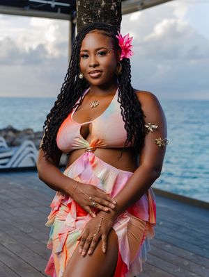 Woman in colorful tie-dye bikini set with pink flower hair accessory posing by the ocean at sunset.