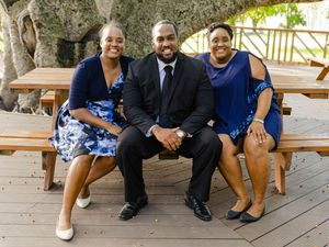 Three people in blue and black formal attire smiling together on a wooden bench outdoors.
