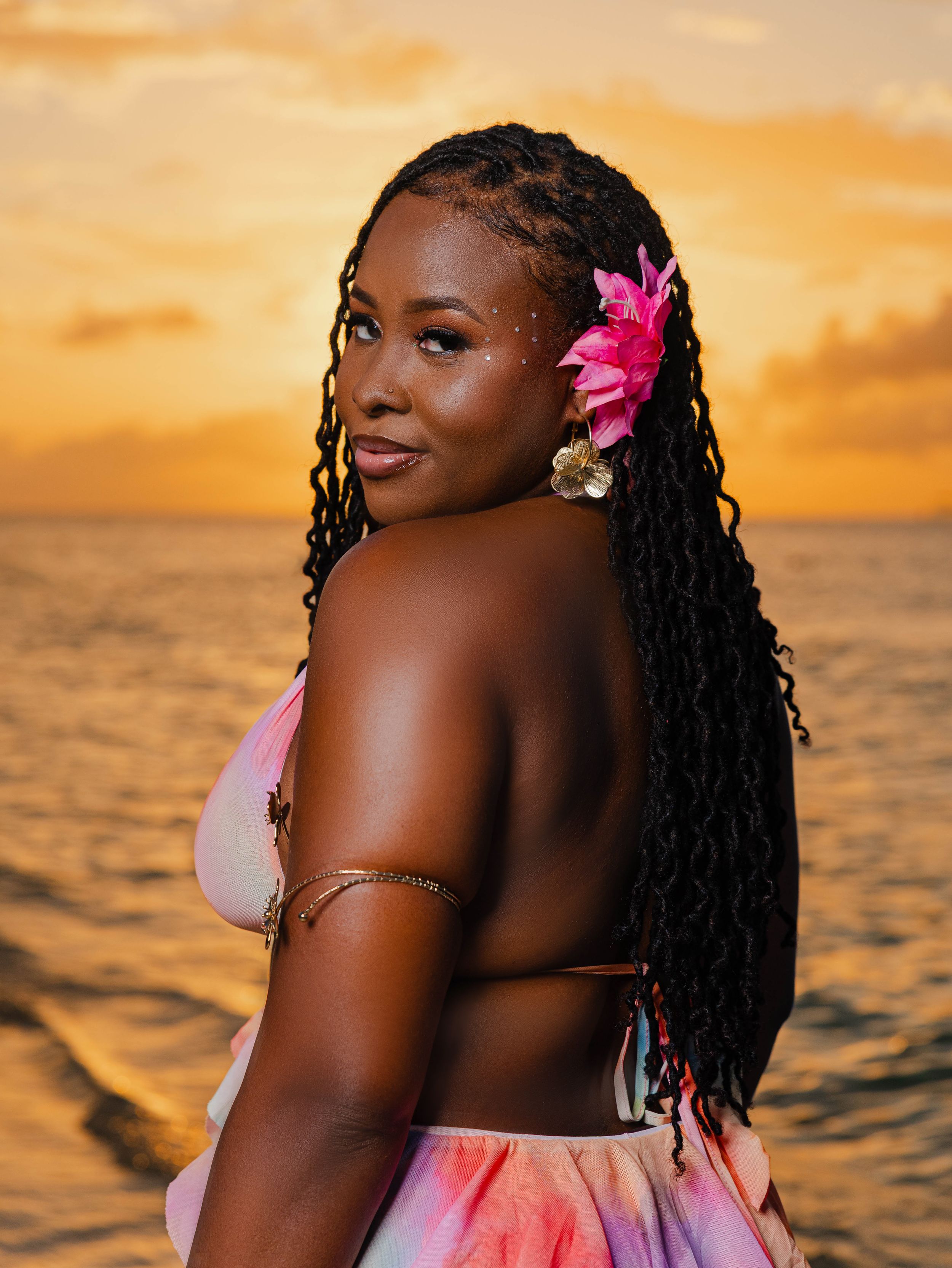 Woman with braids and pink flower at sunset beach in colorful bikini top, golden ocean backdrop.