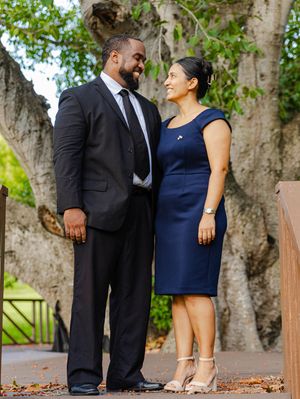 Couple in formal attire sharing a loving gaze while standing together near a large tree on a paved path.