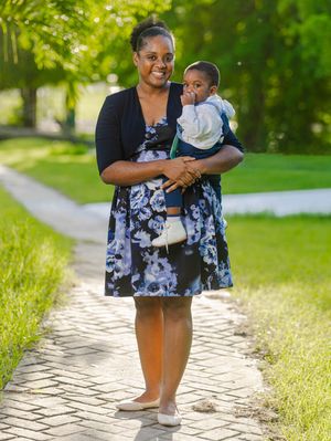Smiling woman in floral dress holds baby boy on a sunlit garden pathway.