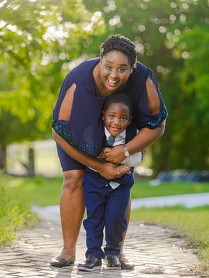 A smiling woman in a navy dress hugs a young boy in a blue suit on a scenic outdoor path.