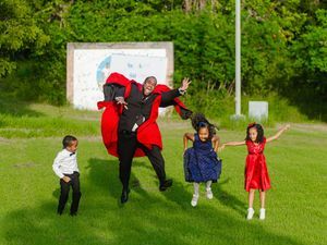 Man in Dracula costume jumping with three children dressed up on green grass outdoors.