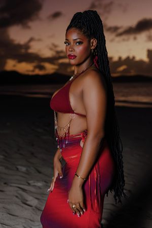 Woman in red bikini top and skirt with long braids poses on a sandy beach at sunset.