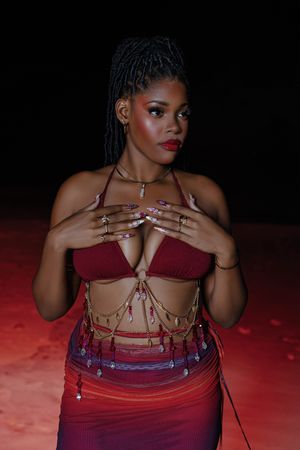 Woman in red halter top and beaded skirt poses confidently against dark background with warm lighting.