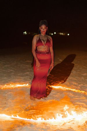 Woman in red two-piece outfit standing in a circle of fire on a beach at night.