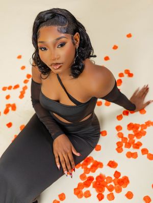 Woman in black off-shoulder dress poses gracefully surrounded by orange rose petals on white background.