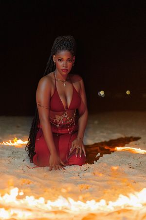 Woman in red outfit kneeling on sandy beach surrounded by fire at night, wearing gold jewelry and braids.