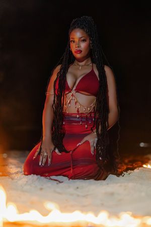 Woman in red outfit with long braids sits on white sand near flames against dark background.