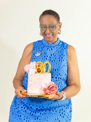 Woman in blue lace dress smiling and holding a pink floral cake with gold 80 topper celebrating her 80th birthday.