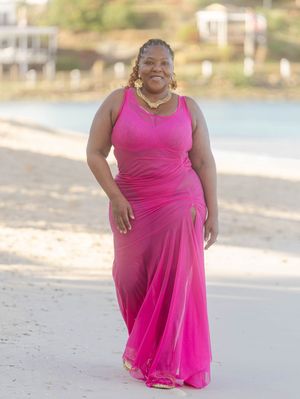 Woman in bright pink maxi dress walking on a sandy beach with coastal buildings in the background.