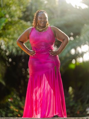 Woman in hot pink mermaid gown posing confidently outdoors with lush green foliage in sunlit background.