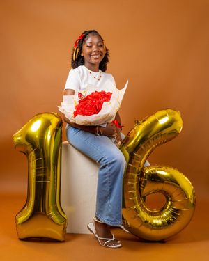 Young girl celebrating her 16th birthday with gold number balloons and red flowers bouquet on brown background.
