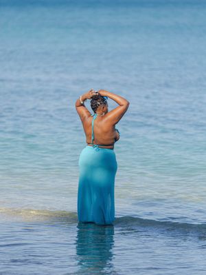Woman in blue sheer skirt and halter top stands in shallow ocean water with hands raised to her hair.
