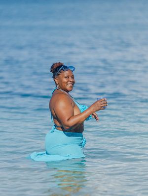 A smiling woman in a blue bikini and sarong stands waist-deep in calm turquoise ocean water on a sunny day.
