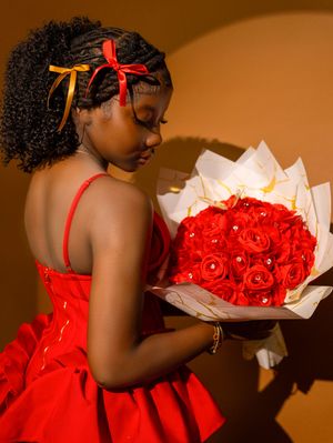 Young girl in red dress holding a bouquet of red roses with white wrapping and gold accents, looking down at flowers.