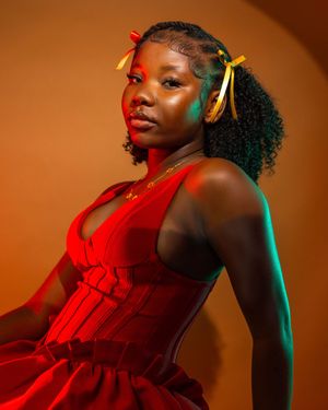 Young Black woman in red dress with yellow hair bows poses confidently against warm orange background.