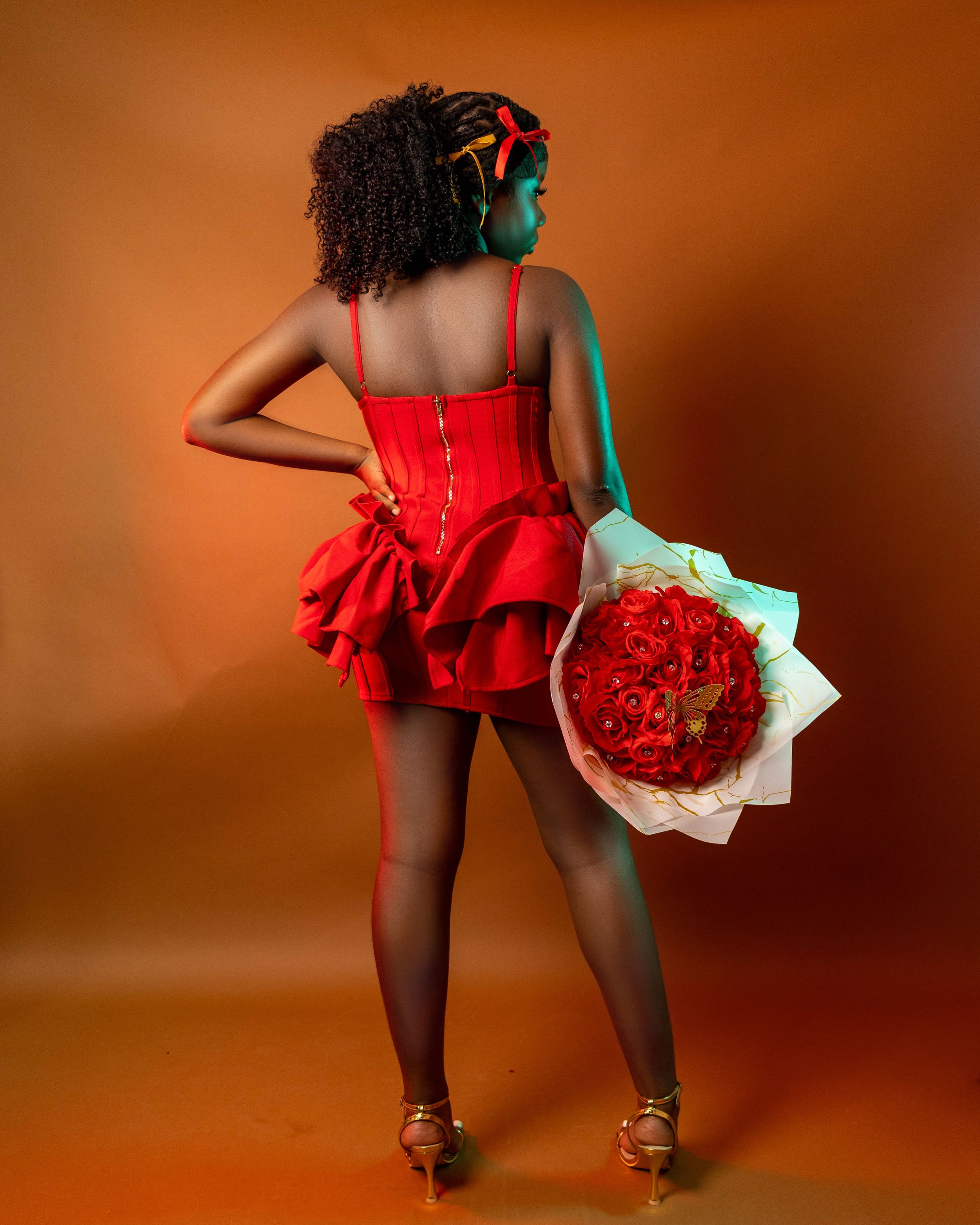 Woman in red ruffled mini dress holding a bouquet of red roses, posing from behind on orange background.