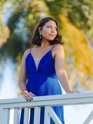 Woman in blue pleated gown stands on white balcony with palm trees in golden hour light.