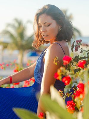 A woman in a blue dress sits among vibrant red flowers with palm trees and golden sunlight in the background.