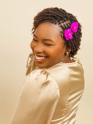 Smiling young Black woman with braided hair adorned with pink flowers, wearing a gold satin top, posing against a beige background.