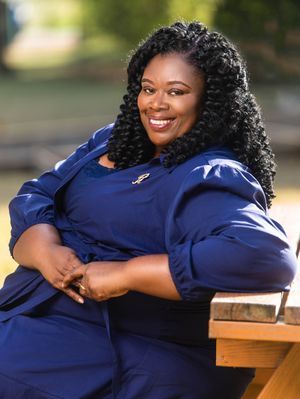 Smiling woman in blue outfit seated outdoors, wearing curly hair and a brooch, posing confidently on a wooden bench.