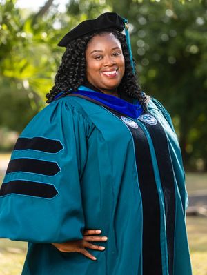 Woman in teal doctoral graduation gown and cap smiling outdoors, celebrating academic achievement.