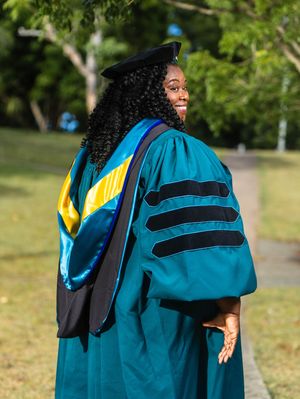 A smiling graduate in teal doctoral gown, black cap, and colorful hood poses outdoors on a green campus.