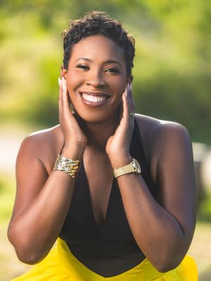 Smiling Black woman with short hair poses outdoors, hands on cheeks, wearing black top and yellow skirt with gold bracelet.