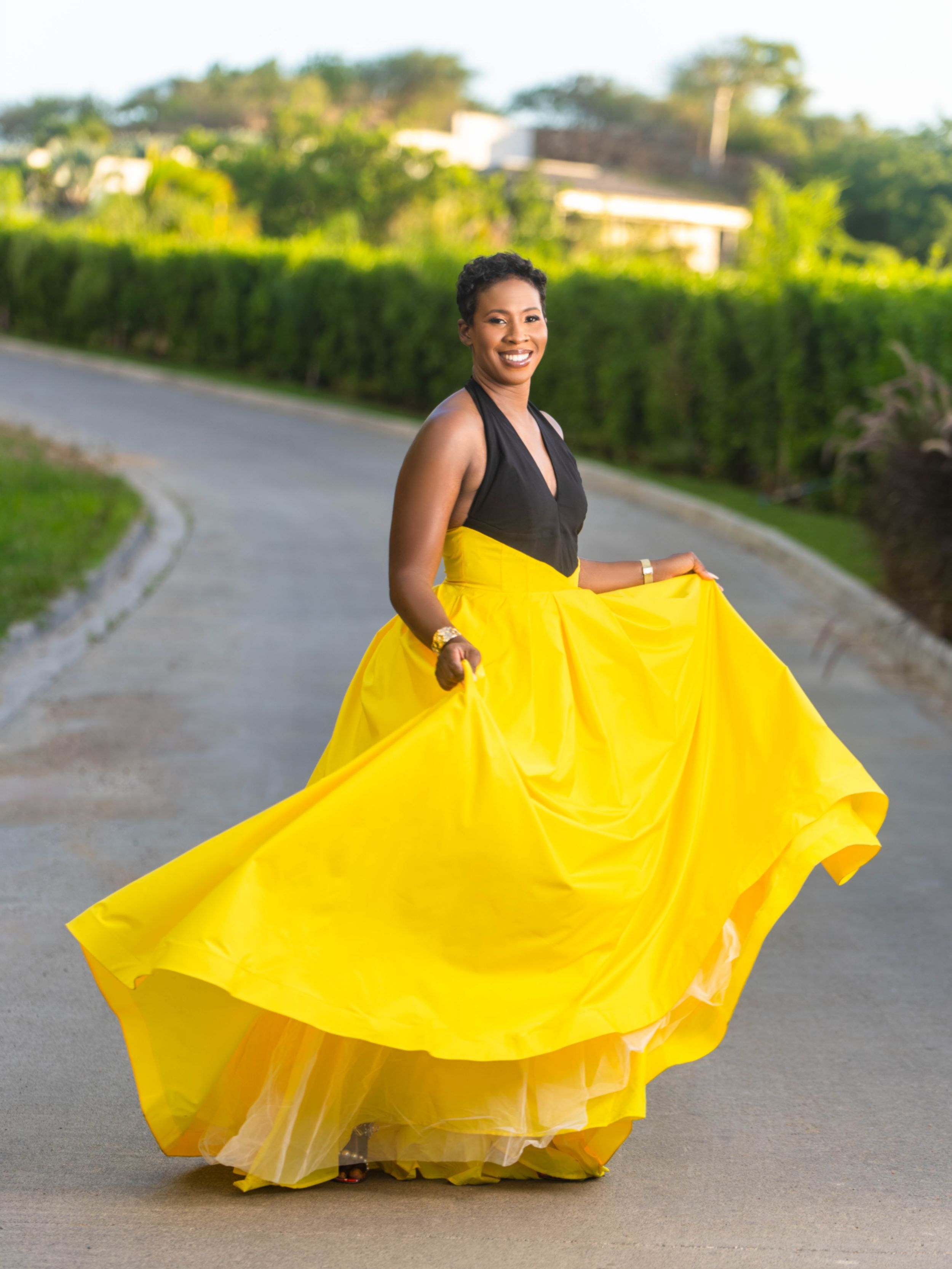 Woman twirling bright yellow maxi skirt on road surrounded by lush greenery.