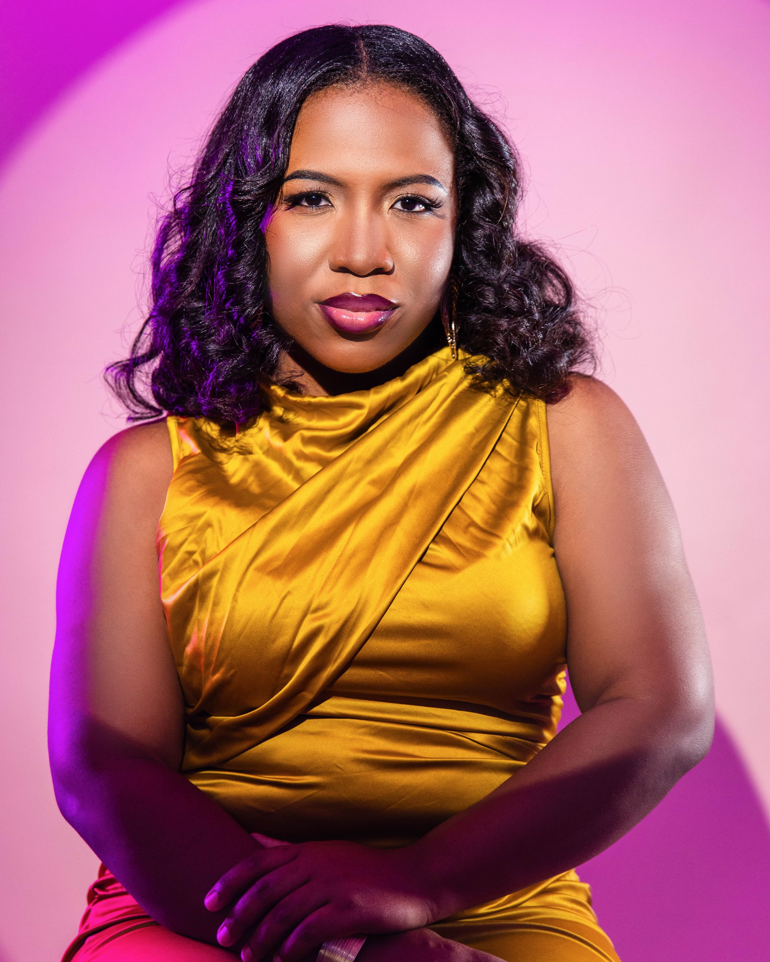 A woman in a gold satin sleeveless dress poses confidently against a vibrant pink background.