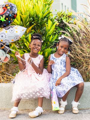 Two young girls in party dresses smile together outdoors, holding balloons and gift bags among green plants.