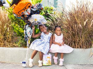 Two young girls in dresses smile holding balloons and a gift bag outdoors near green plants.