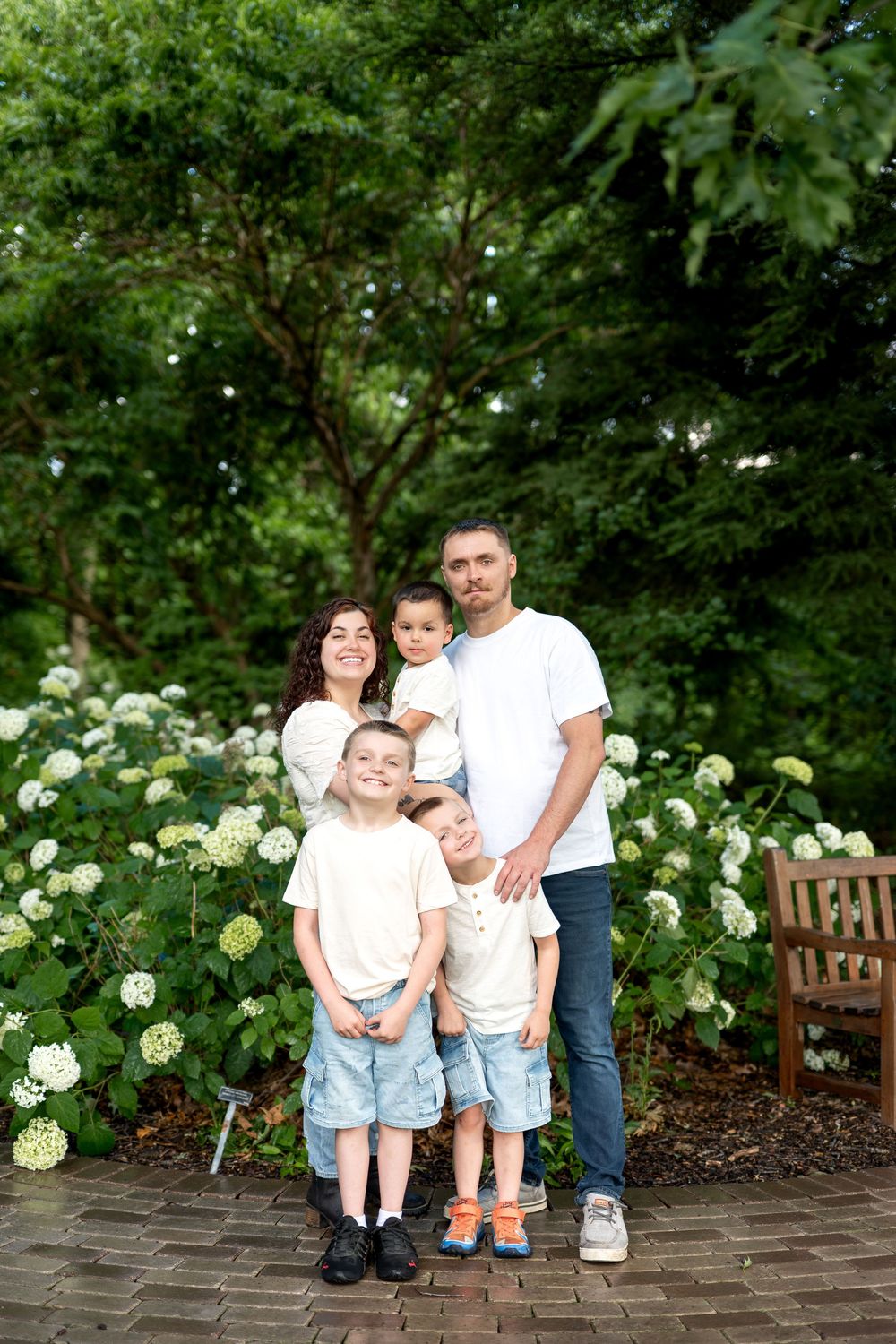 Happy family of five posing in a lush green garden with white hydrangeas, parents and three children wearing white tops.
