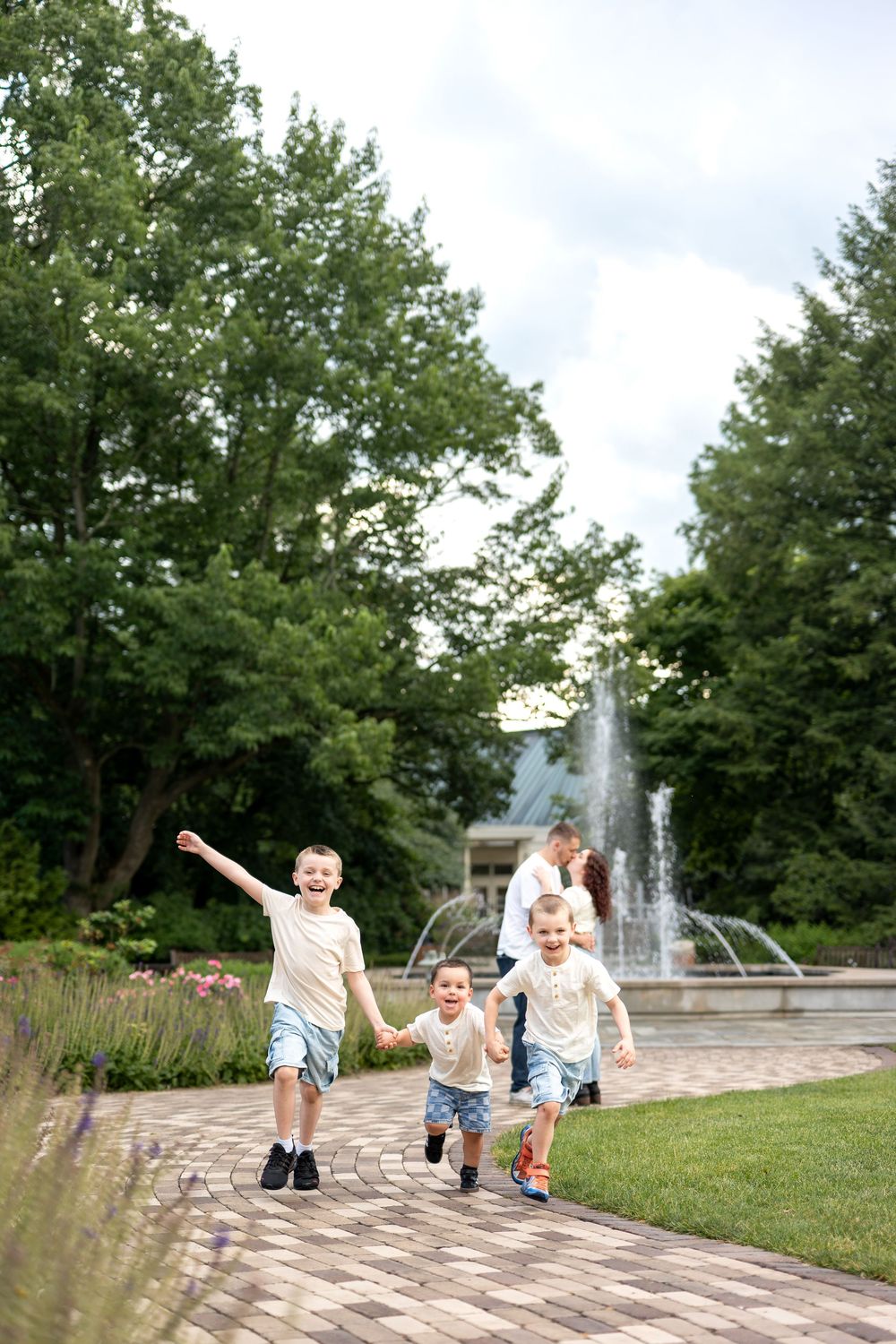 Three children run hand-in-hand along a park path with a fountain and their parents in the background.