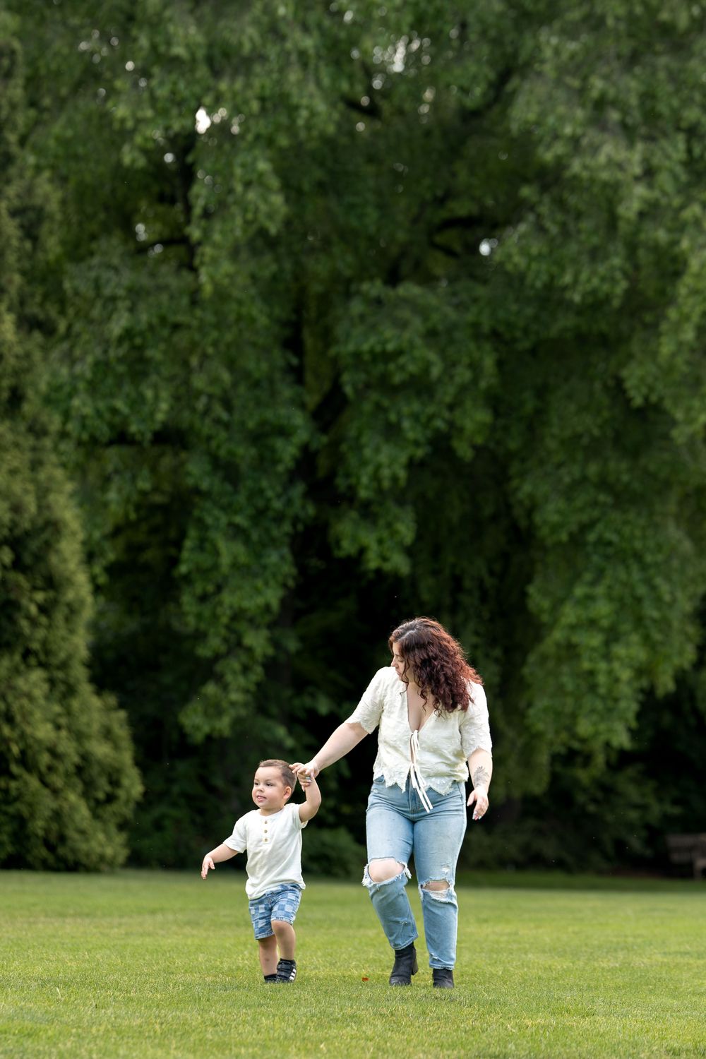 A mother and toddler hold hands while walking on green grass in a lush park on a sunny day.