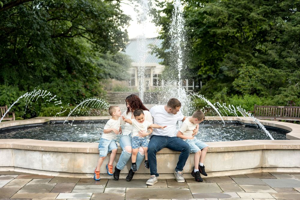 Family of five sitting together at an outdoor fountain surrounded by lush green trees.