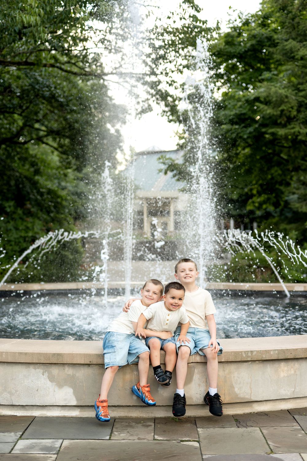 Three boys sitting together on a fountain ledge outdoors, smiling and hugging, surrounded by lush green trees.