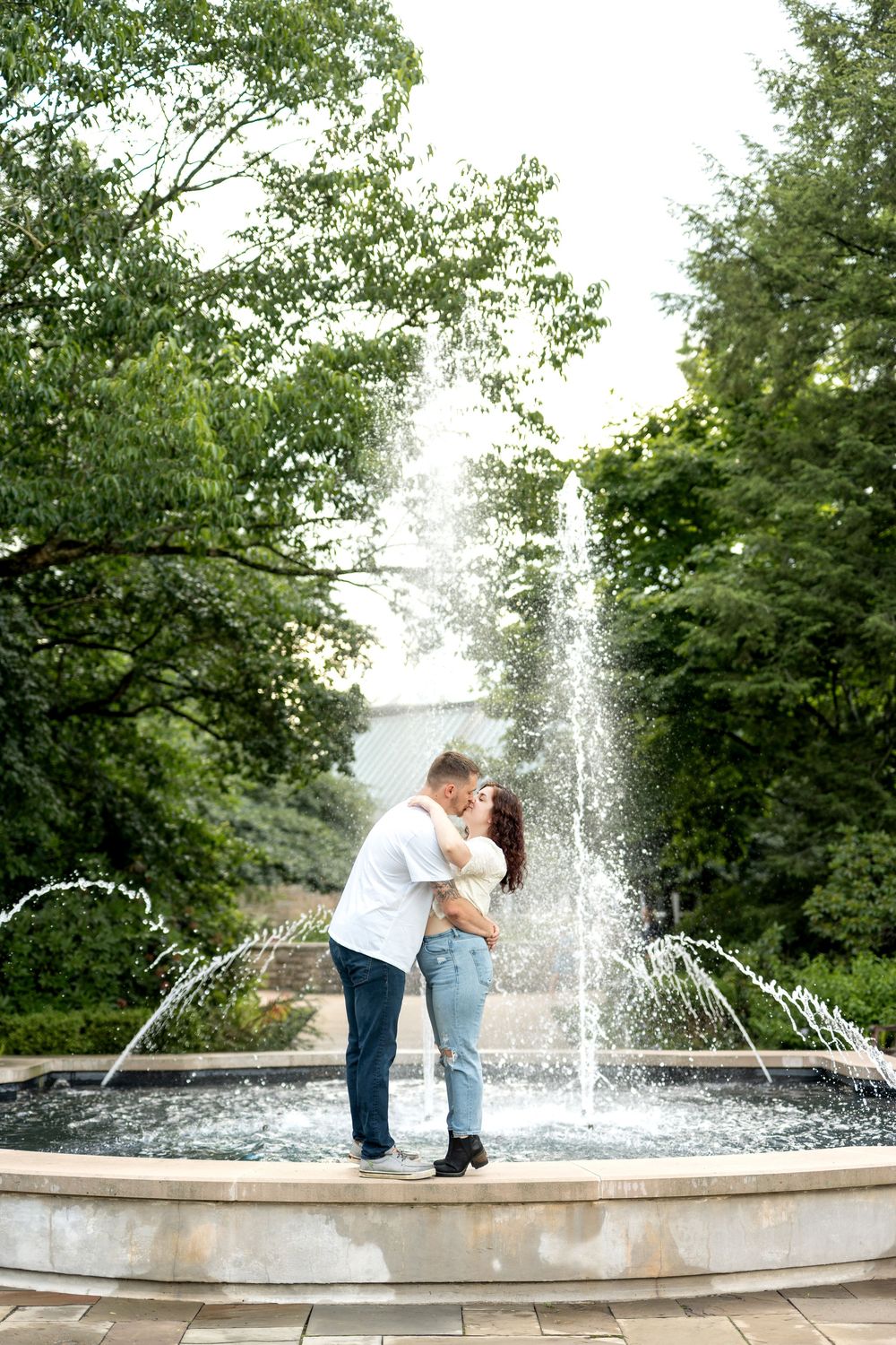 A romantic couple shares a kiss in front of a cascading outdoor fountain in a lush green park.
