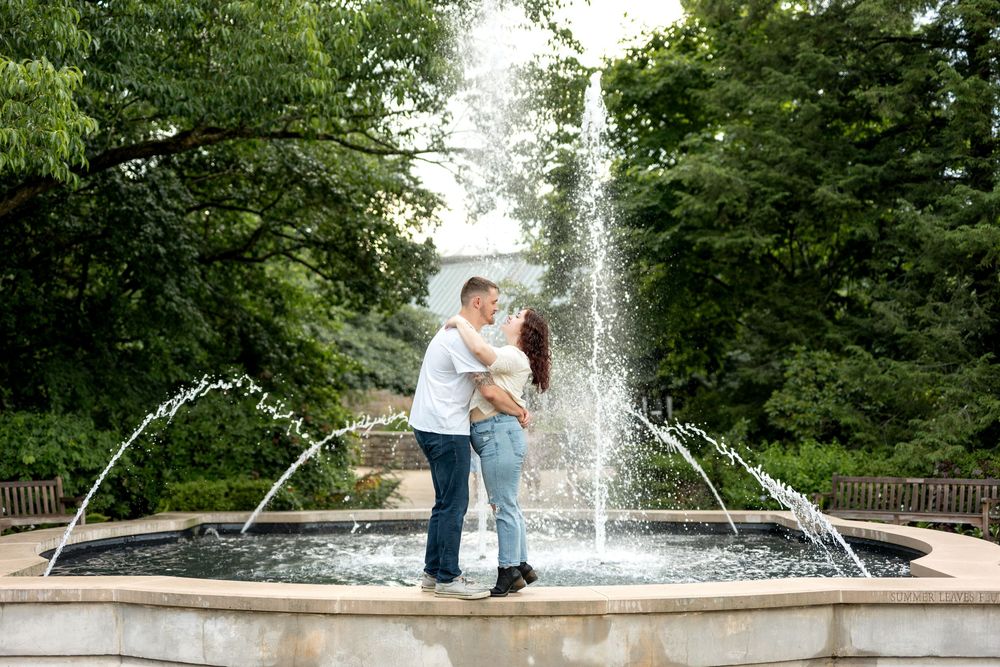 A romantic couple embraces in front of a beautiful fountain surrounded by lush greenery in a park.