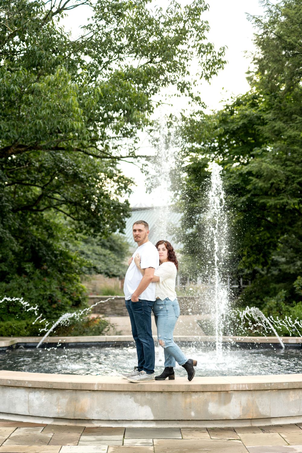 A couple stands back-to-back in front of a beautiful outdoor fountain surrounded by green trees.