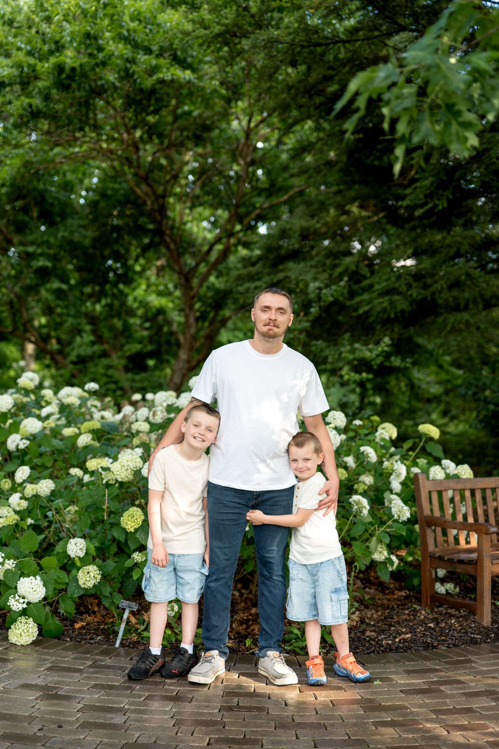 Father posing with two young children in a lush garden with white hydrangeas and green trees.