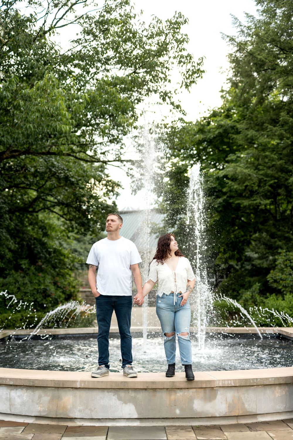 Couple holding hands standing in front of a fountain surrounded by lush green trees in a park setting.