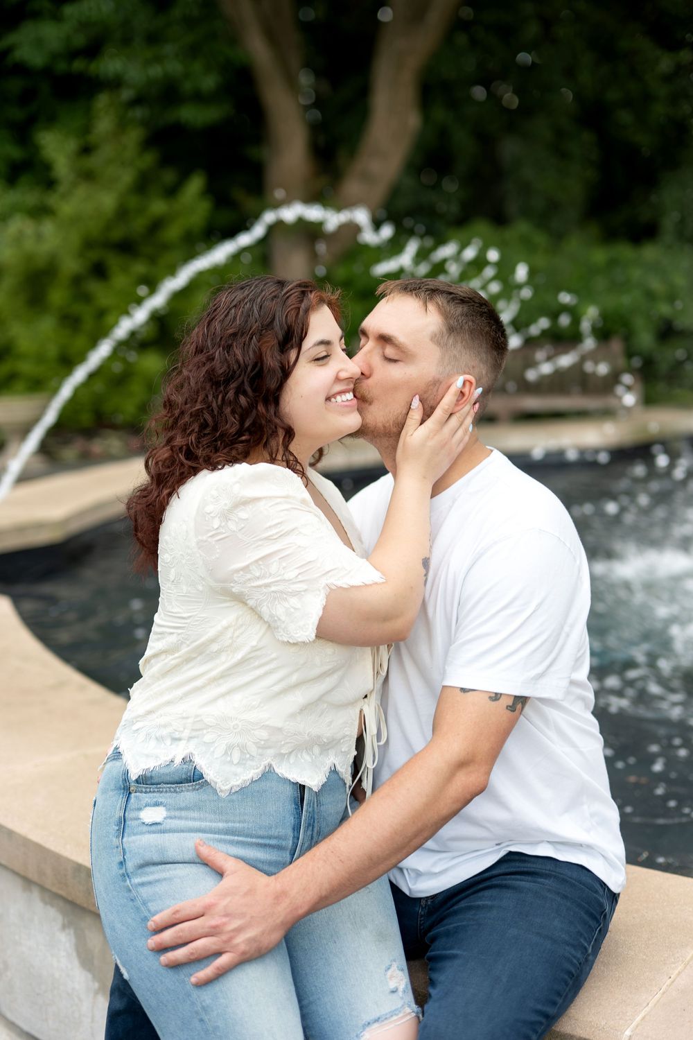 Couple sharing a romantic kiss by a fountain outdoors, woman in white lace top and jeans, man in white t-shirt.