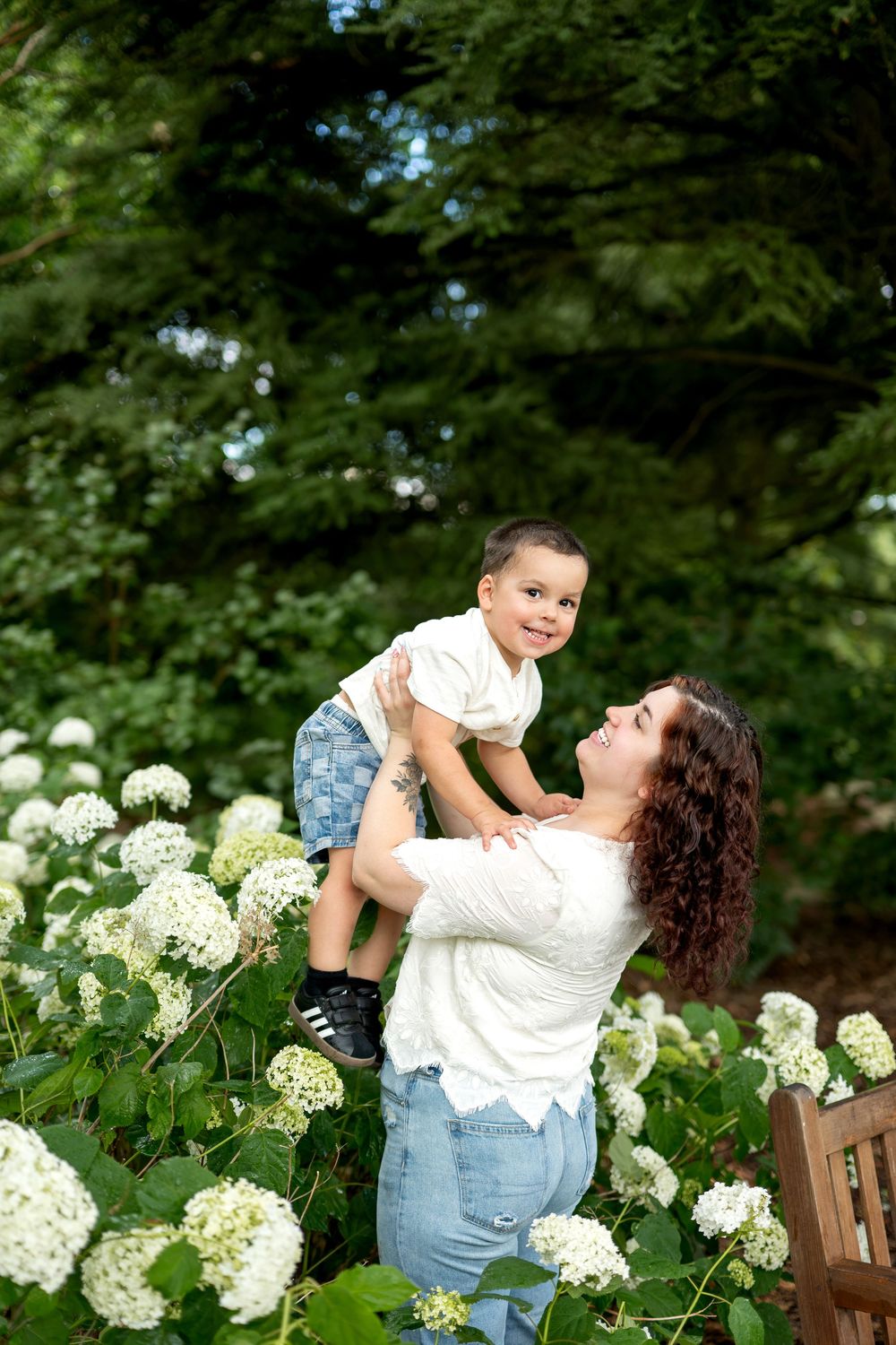 A mother lifts her smiling toddler among white hydrangeas in a lush green garden, both wearing casual white tops.