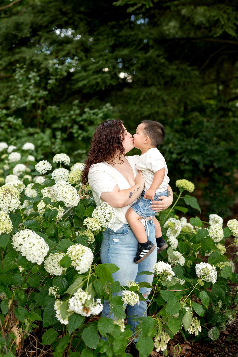 Mother kissing toddler son while standing among white hydrangea bushes in a lush green garden.